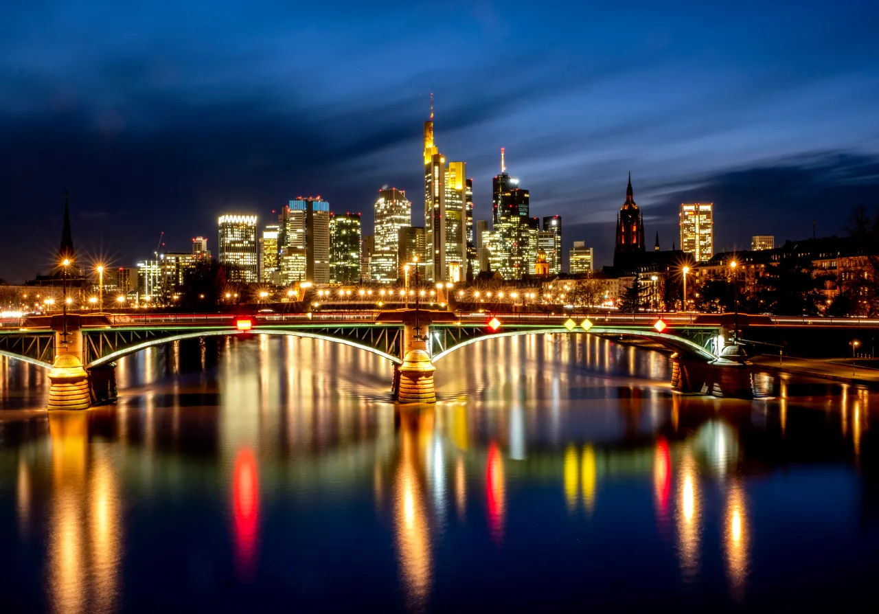 Nächtliche Stadtsilhouette mit beleuchteten Wolkenkratzern und einer Brücke, die sich in einem Fluss spiegelt, in dunklen Blau- und Goldtönen